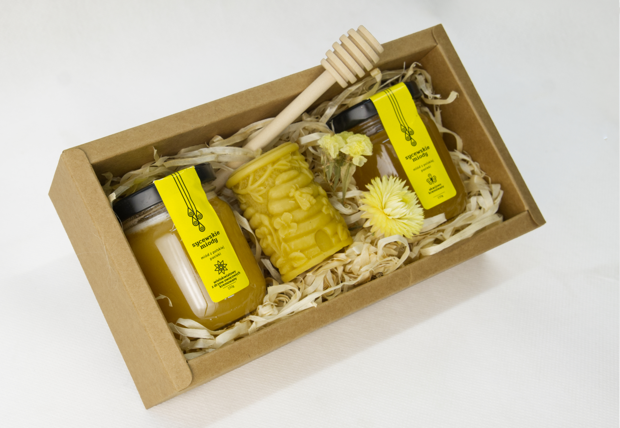 Dwa małe słoiki miodu ze świeczką z wosku pszczelego oraz drewnianym miodownikiem w kratowym bocxie.